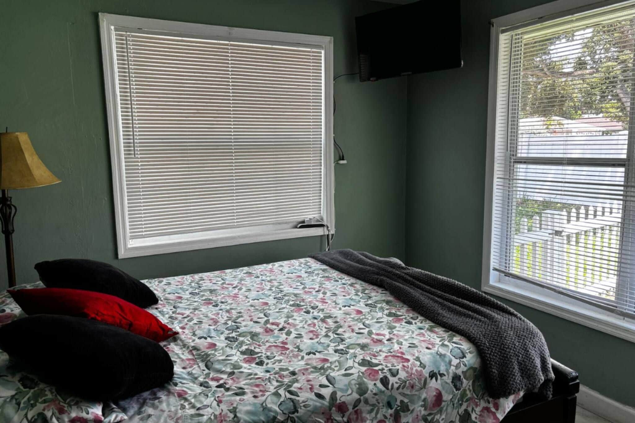 A bedroom with a bed, a window with blinds, a lamp, and a television on the wall.