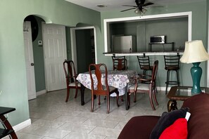 Zona pranzo con tavolo e sedie, ventilatore a soffitto e angolo cottura con elettrodomestici.