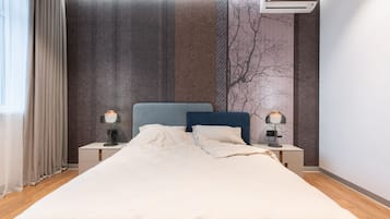 A neatly made bed with a headboard, two bedside lamps, a wall-mounted air conditioner, and a window with curtains.
