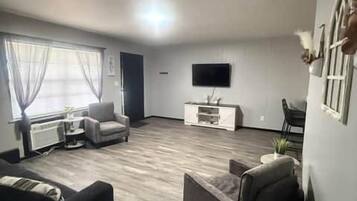 Una sala de estar con televisor de pantalla plana, sofá, mesa de centro y ventana con cortinas.