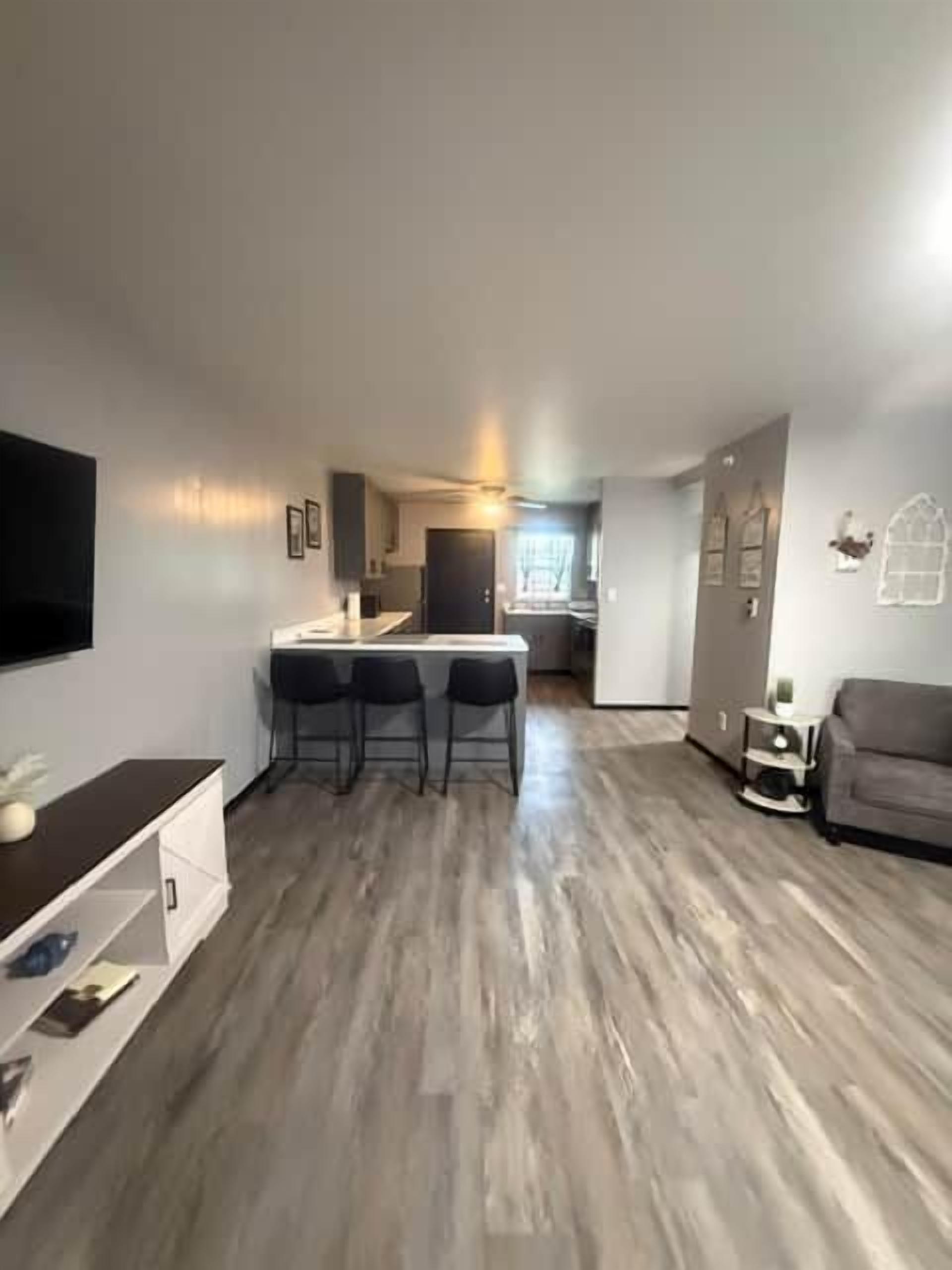 A modern living room with a flat-screen TV, a dining area with a bar stool, and a kitchen in the background.