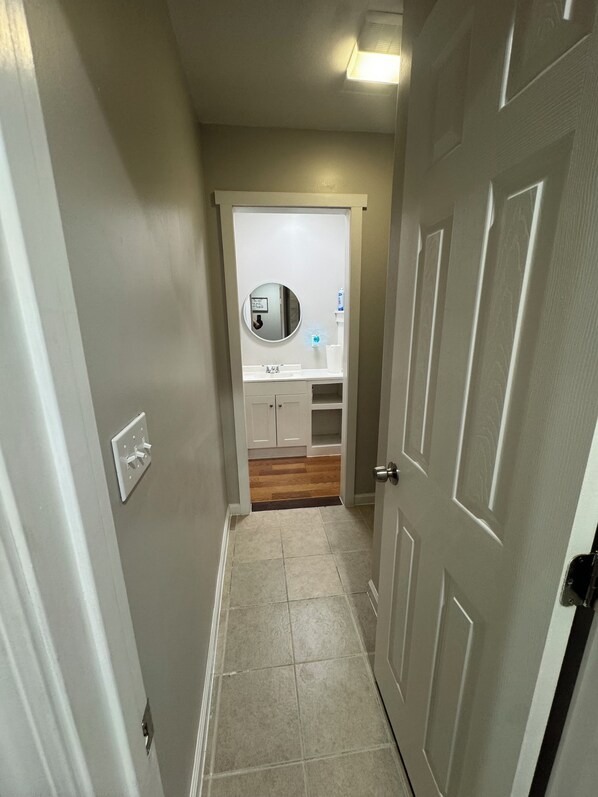 A narrow hallway with a door on the right, a light switch on the left, and a bathroom visible at the end with a mirror, sink, and toilet.