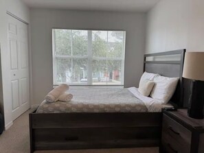 Quarto com cama, mesa de cabeceira, abajur e janela com persianas.
