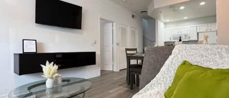A modern living room with a glass coffee table, a sofa with a green pillow, a flat-screen TV mounted on the wall, and a kitchen area in the background.