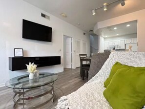 A modern living room with a glass coffee table, a sofa with a green pillow, a flat-screen TV mounted on the wall, and a kitchen area in the background. - Casa familiar de 3 habitaciones con piscina, cerca de Disney (Kissimmee)