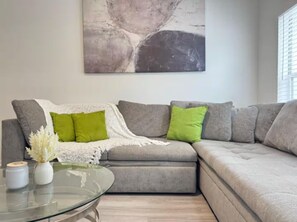 Uma sala de estar moderna com um sofá seccional cinza, duas almofadas verdes, um cobertor branco e uma mesa de centro de vidro com dois recipientes brancos.