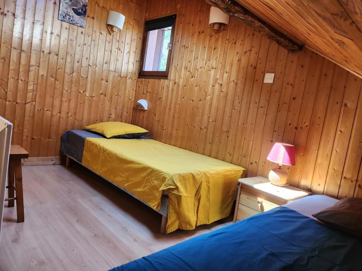 Una habitación con paredes de madera y techo inclinado, con una cama con colcha amarilla, una mesita de noche con lámpara y una ventana.