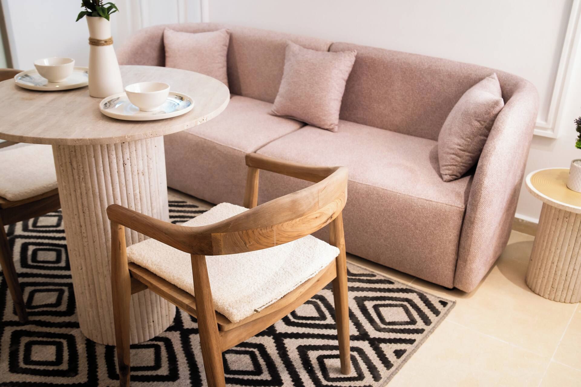 A living room with a beige sofa, a round wooden table, a single wooden chair, and a patterned rug.