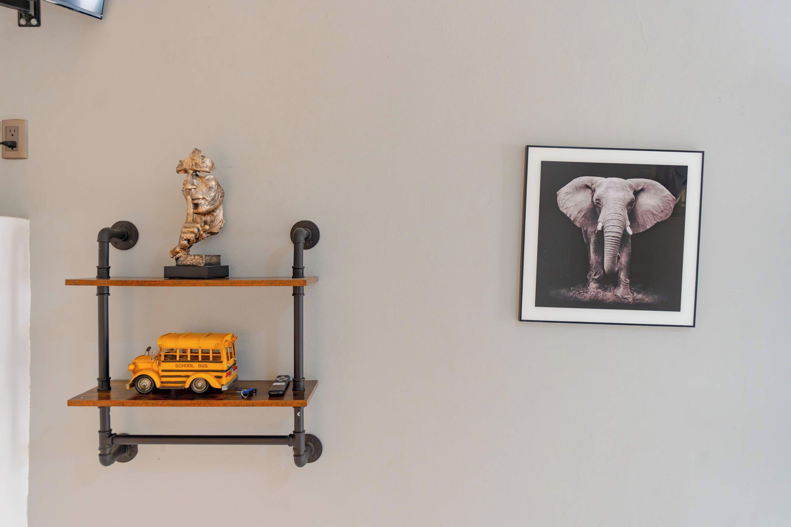 Una pared con un estante que sostiene una escultura, un modelo de autobús escolar y un cuadro con una imagen de elefante.