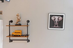 Un mur sur lequel est fixée une étagère, et sur laquelle sont posés une sculpture, une maquette de bus scolaire et un tableau encadré représentant un éléphant.