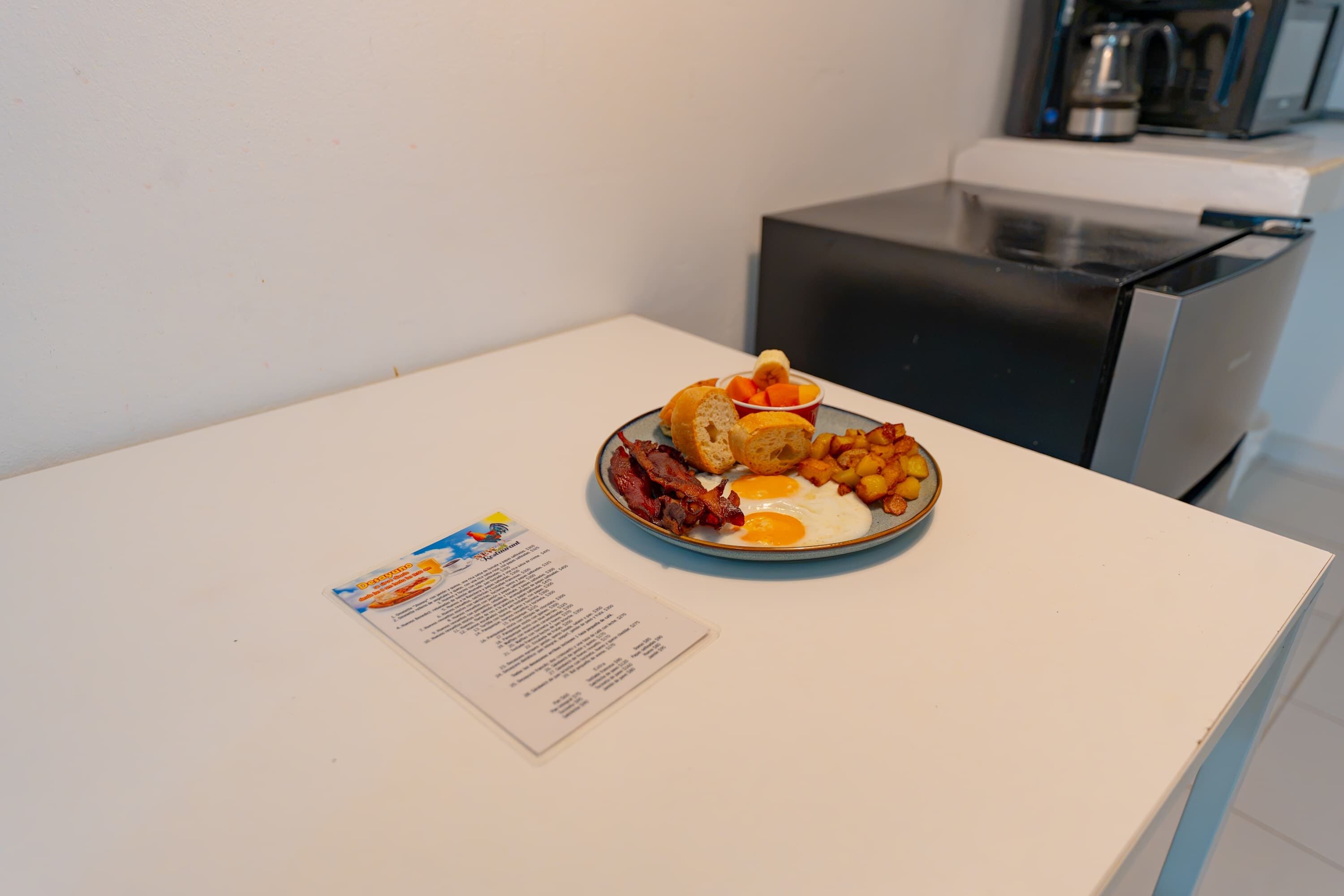 Une assiette de petit-déjeuner avec des œufs, du bacon, des toasts et des fruits, posée sur une table blanche, avec un menu à proximité.