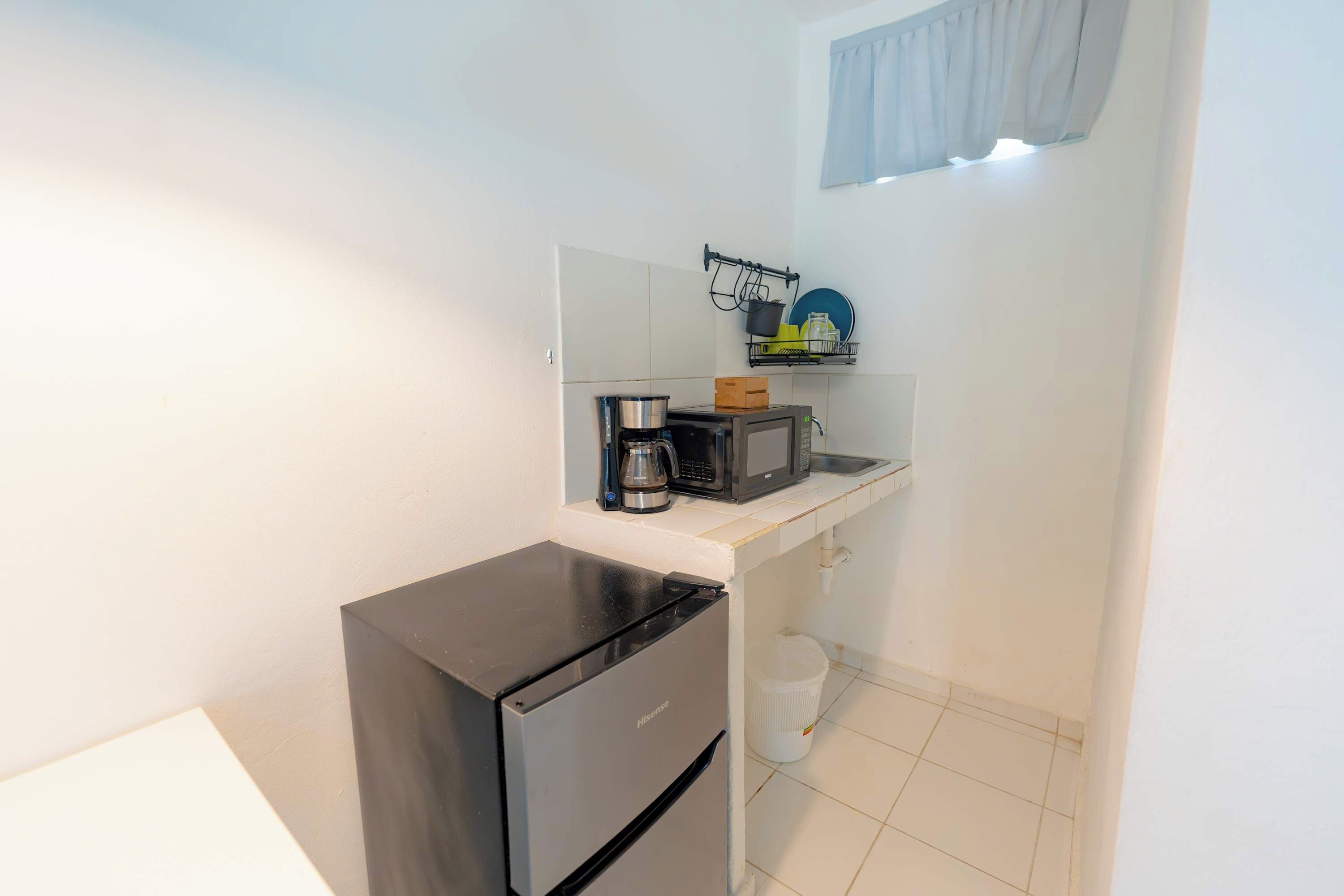 Una kitchenette compacta con refrigerador, microondas, cafetera y un pequeño estante con vajilla.