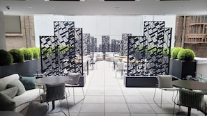Uma área de estar moderna com sofás, mesas e cadeiras, separada por divisórias estampadas em preto e branco.