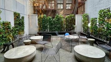 An outdoor seating area with round tables and metal chairs, surrounded by greenery and brick walls.