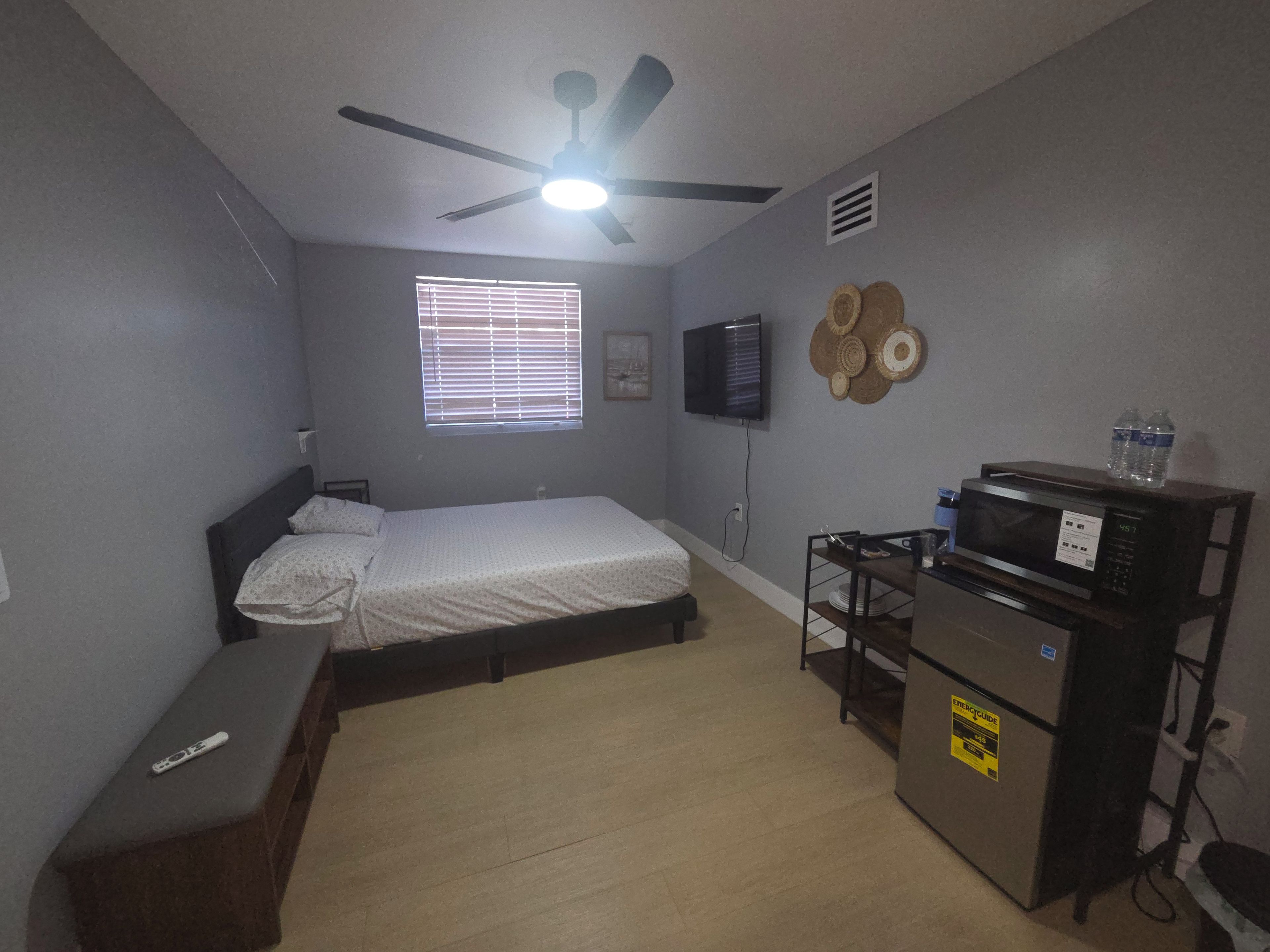 Habitación compacta con cama, ventilador de techo, televisor, microondas y una pequeña mesa con una botella de agua.