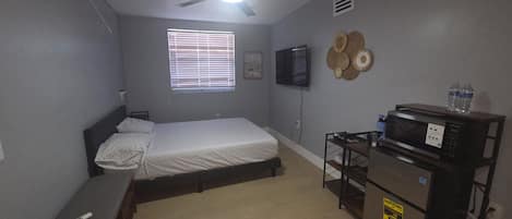 Habitación compacta con cama, ventilador de techo, televisor, microondas y una pequeña mesa con una botella de agua.