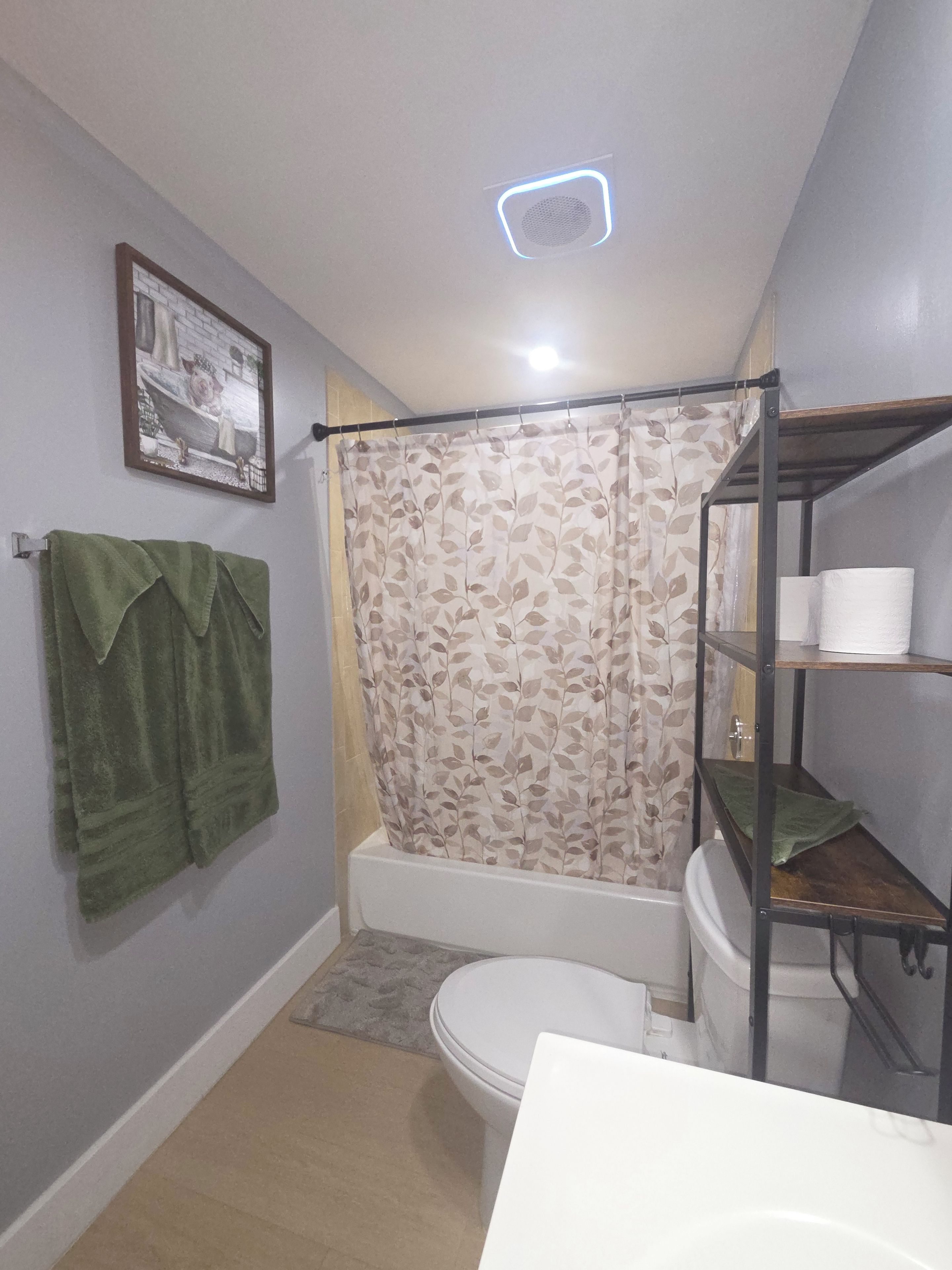 Un baño con inodoro, bañera, cortina de ducha, toallero con toallas verdes y repisa con papel higiénico.
