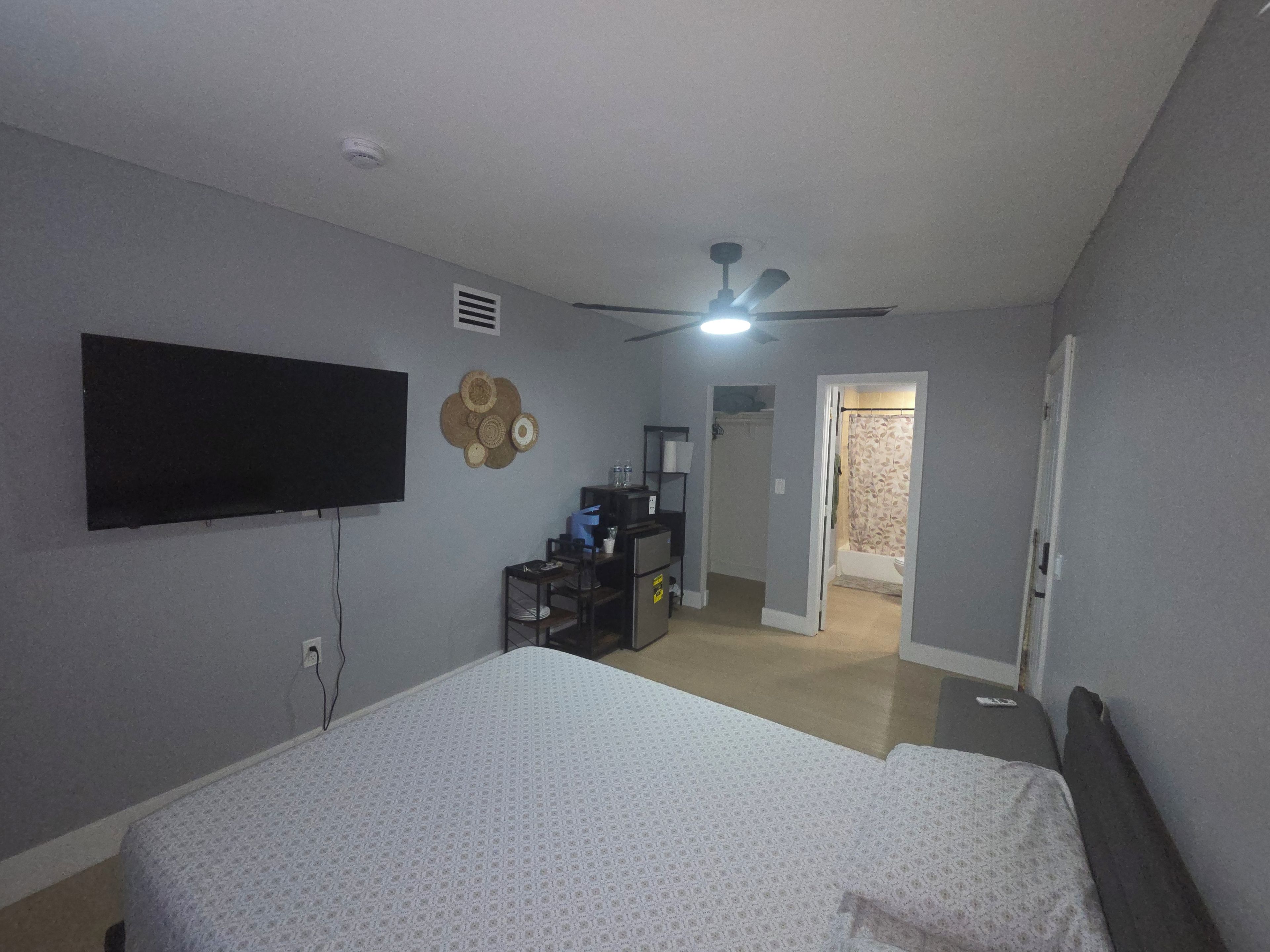 Un dormitorio con cama, ventilador de techo, televisión montada en la pared y un baño a la vista a través de una puerta abierta.