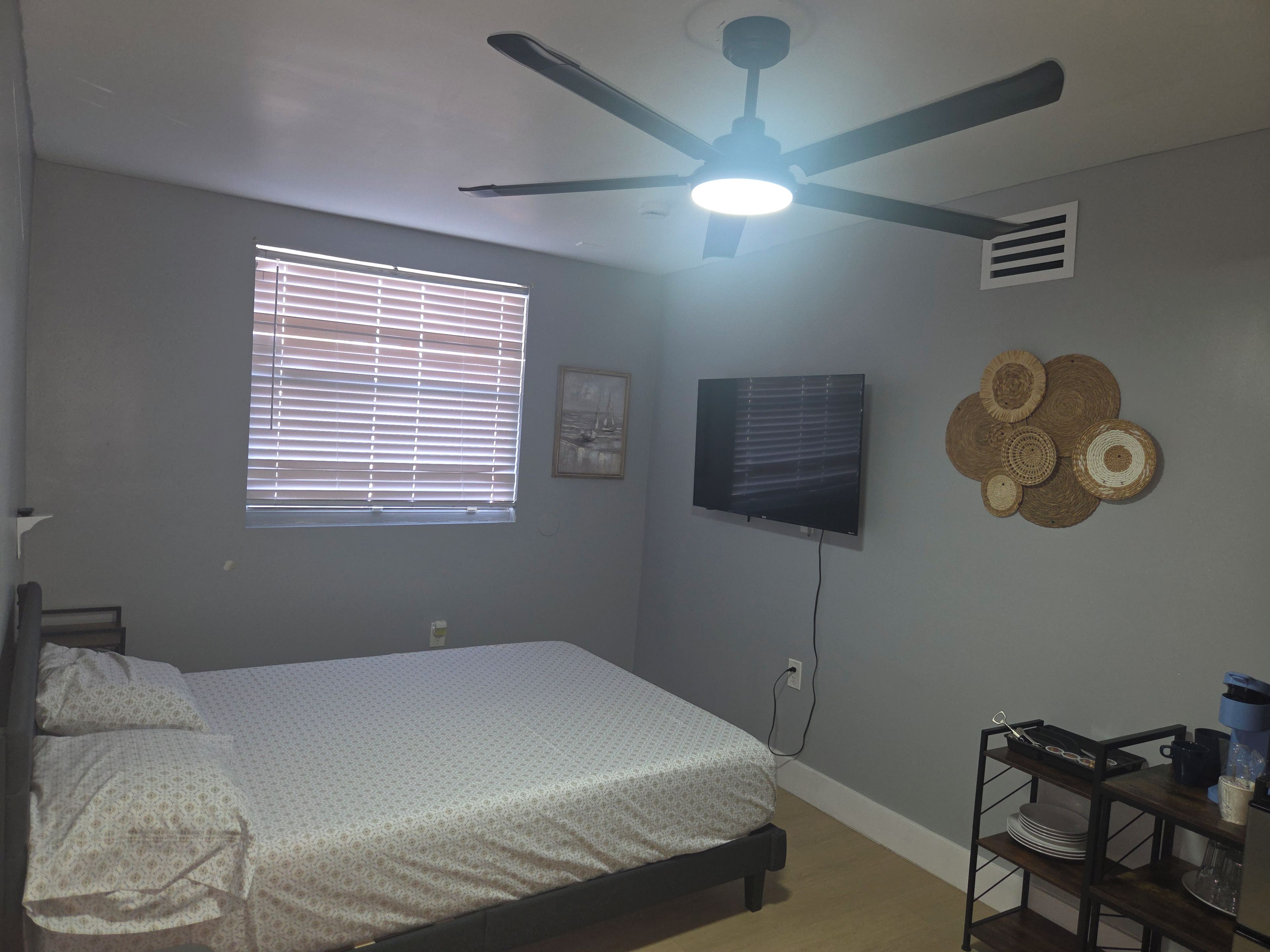 Un dormitorio con una cama, un ventilador de techo, un televisor, una ventana con persianas y una estantería en la pared con vajilla.