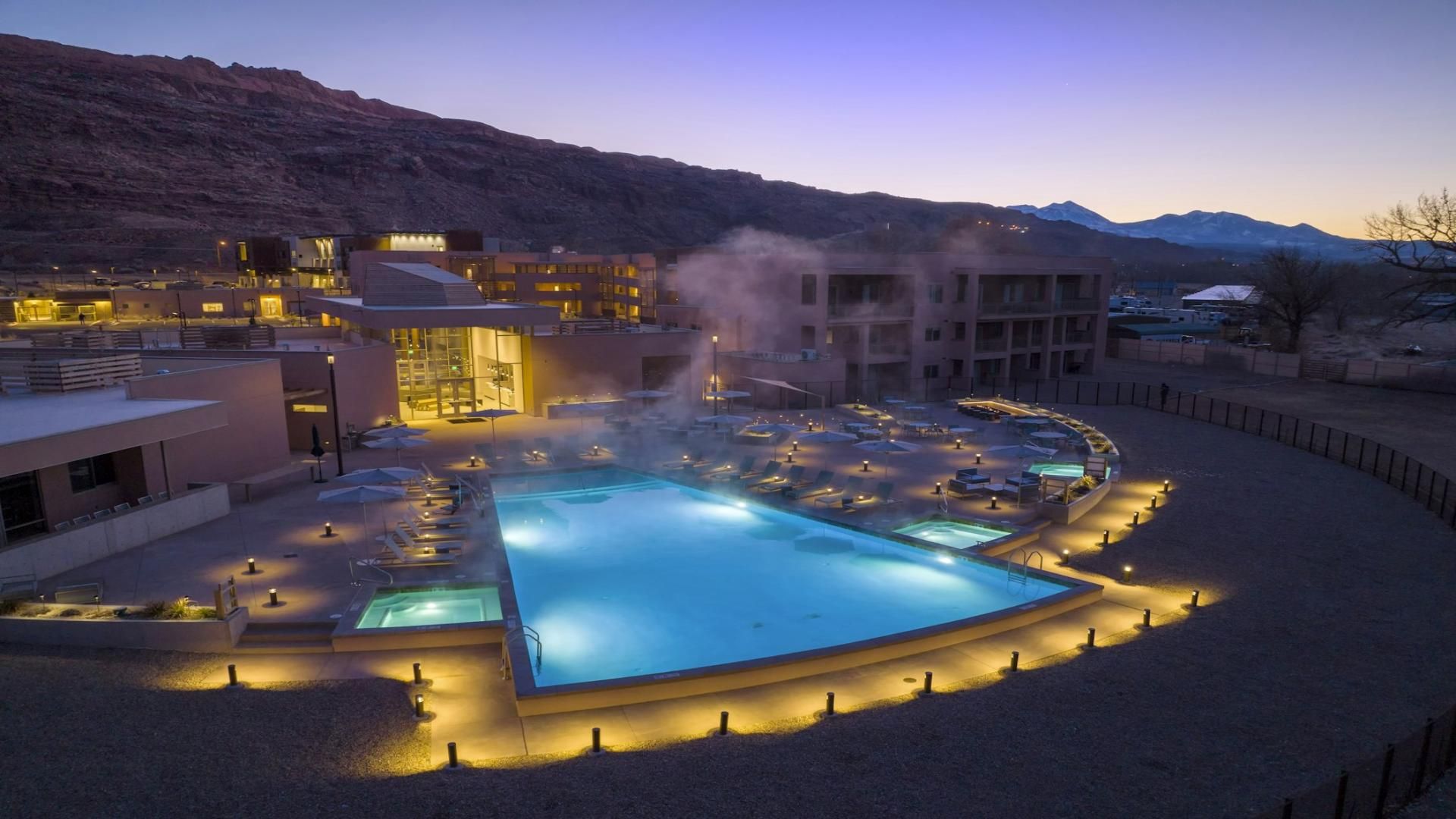 Un hotel con una amplia piscina exterior, zonas de descanso y montañas al fondo.