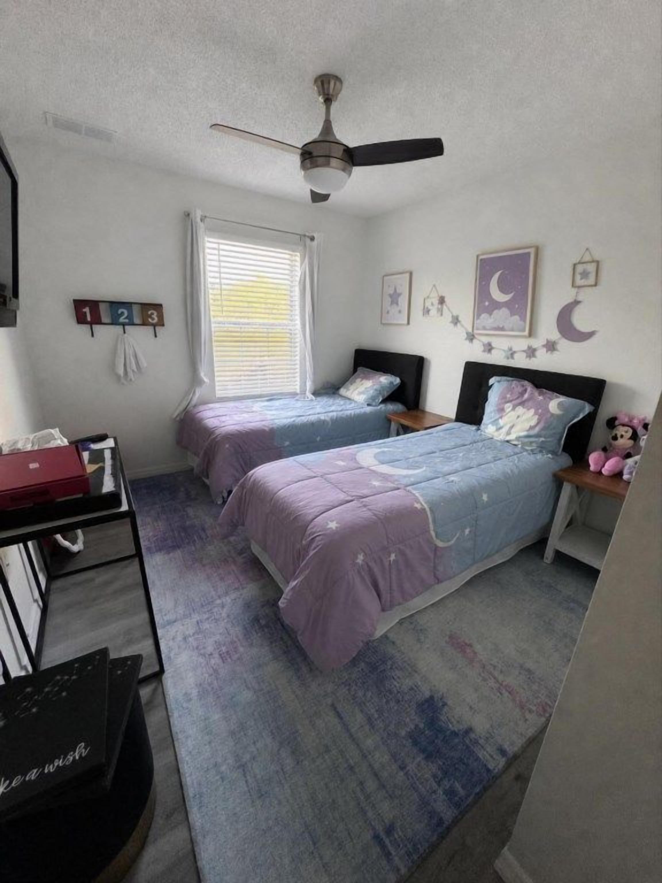 Une chambre avec deux lits, un ventilateur de plafond, une fenêtre avec des stores et une horloge murale.