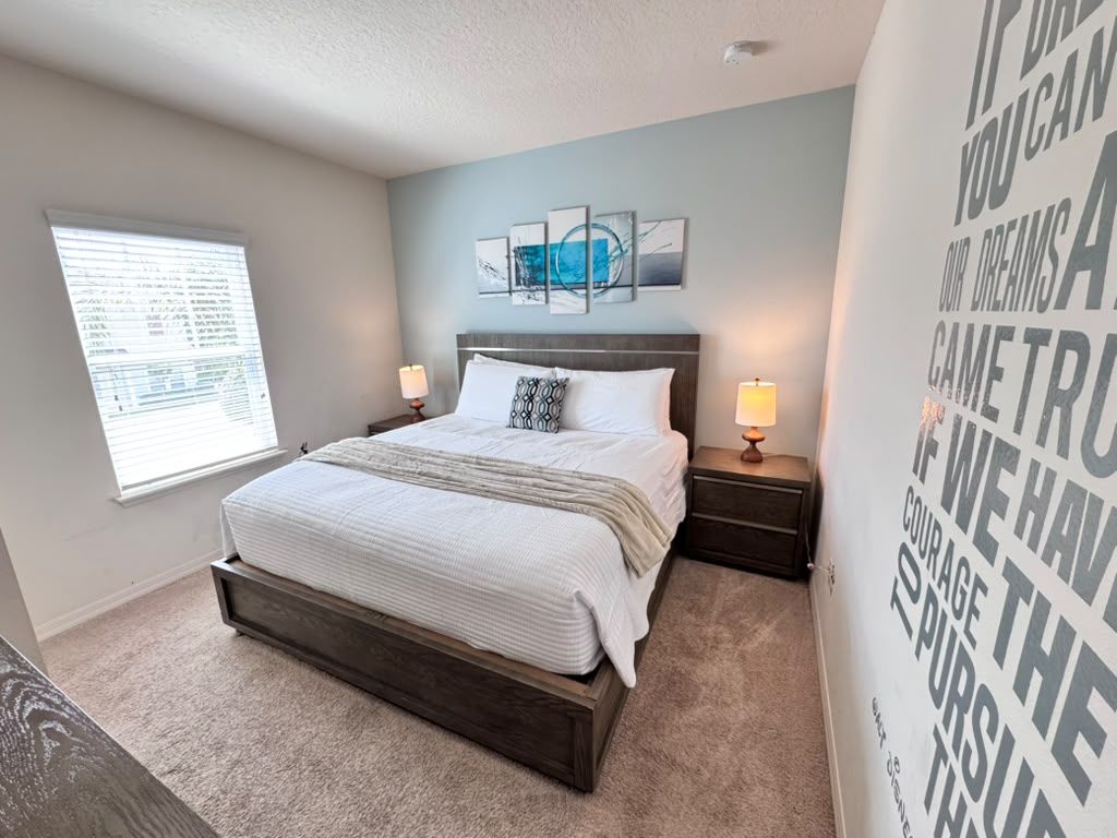 A bedroom with a bed, bedside tables, a window with blinds, and a motivational quote on the wall.