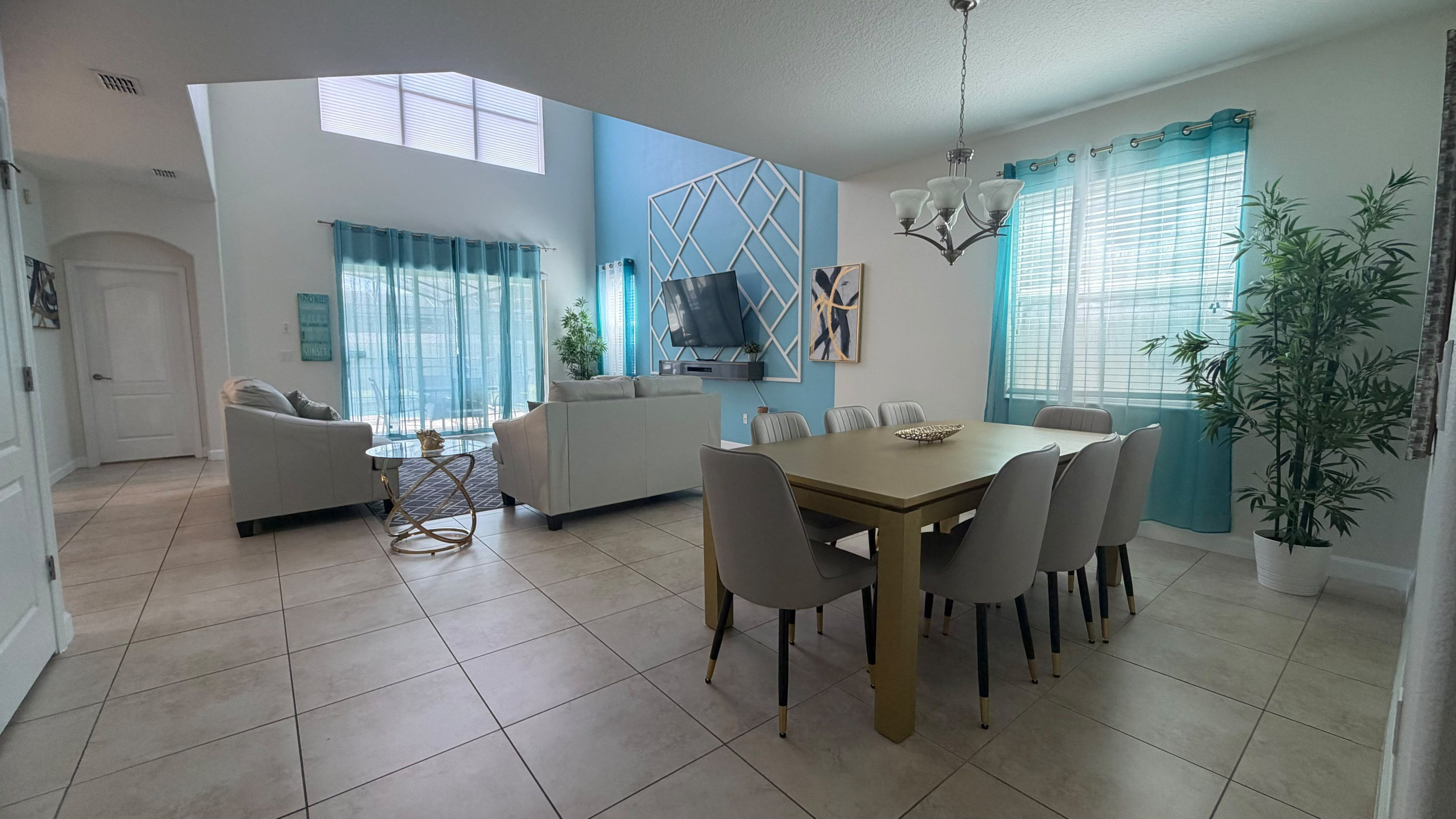 A spacious living room with a dining area, a TV mounted on a wall, and a large window with curtains.