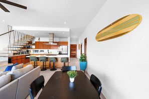 Eine moderne Küche mit Holzschränken, einer zentralen Kochinsel inklusive Barhocker und einem Essbereich mit dunklem Holztisch und Stühlen.