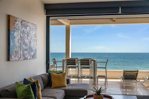 A modern living room with a large window overlooking the sea, a sofa with cushions, and a painting on the wall. - Oceanfront Cove Condo | Ocean-View Terrace & Pool (El Pescadero)