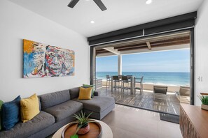 A modern living room with a gray sofa, a colorful abstract painting, and a view of the ocean through large glass doors. - Oceanfront Cove Condo | Ocean-View Terrace & Pool (El Pescadero)