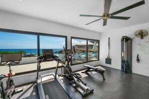 Ein Fitnessraum mit Trainingsgeräten, großen Fenstern mit Meerblick und einem Deckenventilator.