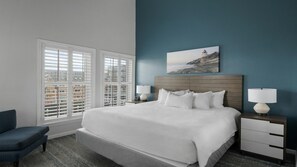 A hotel room with a large bed, two bedside tables with lamps, a chair, and a view of buildings through the windows.
