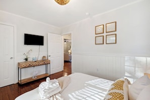 A hotel room with a bed, a TV mounted on the wall, a console table with a fan, and a door leading to another room. - Quaint Cottage-Great Location with Hot Tub! (Mount Pleasant)