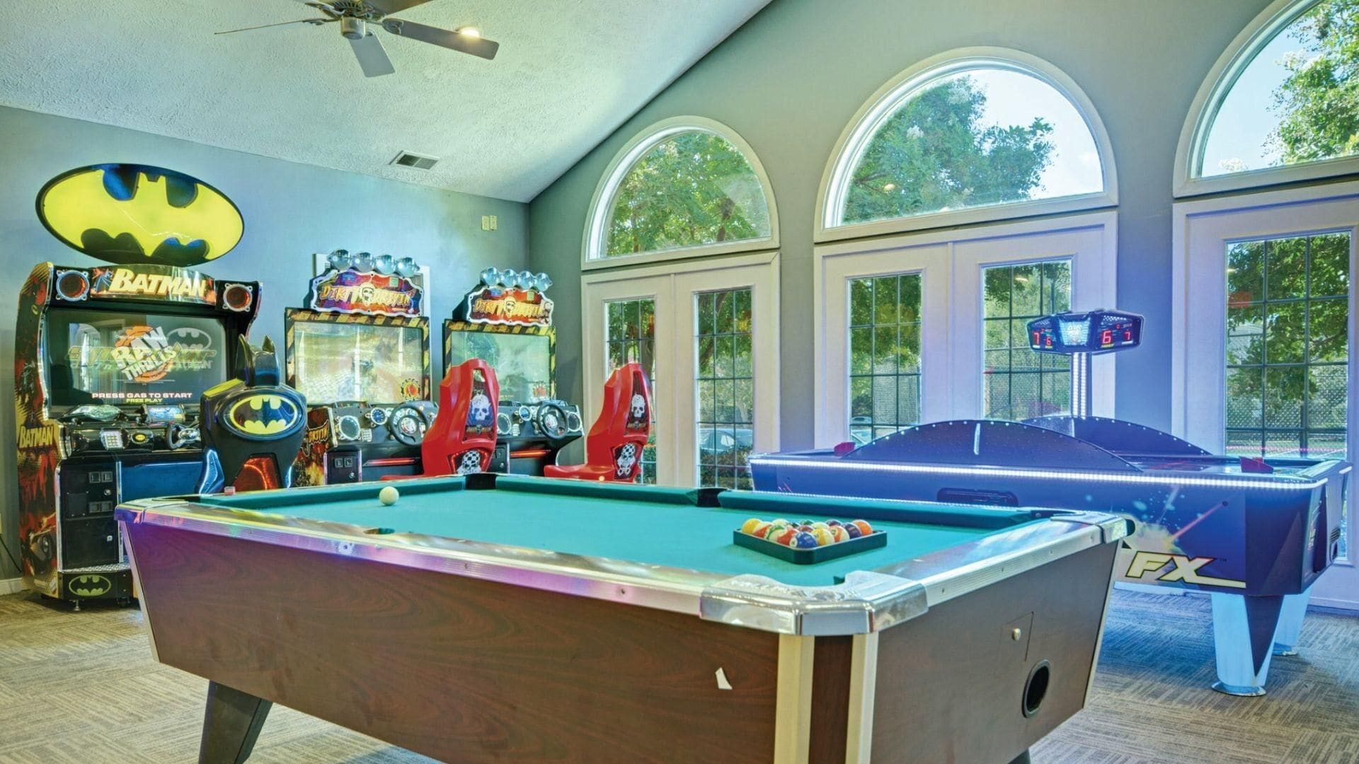 Une table de billard dans une pièce où se trouvent des jeux d’arcade et de grandes fenêtres.