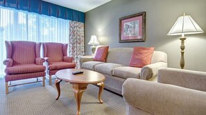 Une chambre d’hôtel avec un canapé, deux fauteuils, une table basse, une lampe, un cadre photo et une fenêtre avec des rideaux.