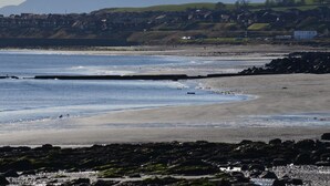 Une plage côtière avec un premier plan rocheux, une rive sablonneuse et une vue lointaine sur des maisons et des collines.