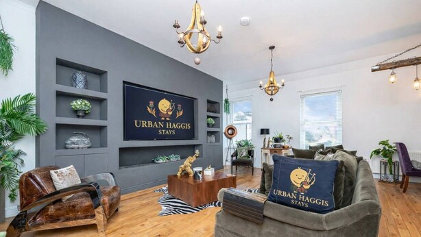 Un salon moderne avec un canapé gris, une table basse en bois et un téléviseur fixé au mur qui affiche le message « URBAN HAGGIS STAYS ».