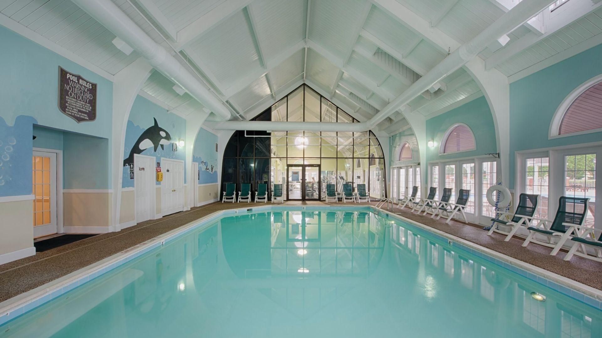 An indoor swimming pool with clear water, lounge chairs, and a pool rules sign on the wall.