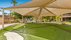 Un campo de golf con una estructura tipo quiosco, una piscina y un edificio con balcones al fondo.