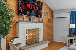 A living room with a fireplace, a mounted television displaying a Netflix interface, and a wall-mounted air conditioning unit. - Private Heated Pool, Hot Tub, Sauna, Fire Pit (Pocono Summit)