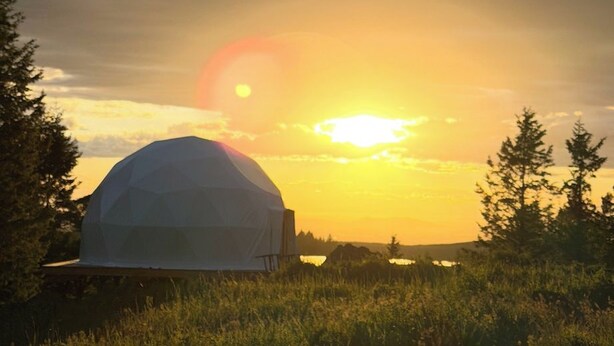 Estrutura em forma de cúpula geodésica em um campo gramado, com o pôr do sol ao fundo.