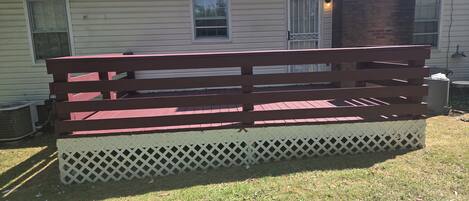 Un banc en bois doté d’une base ajourée, placé devant une maison ornée d’un pilier en briques et d’une fenêtre avec une grille en métal.