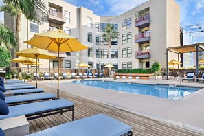 Zona de piscina con sillas de descanso y sombrillas, rodeada por un edificio de varios pisos con balcones.