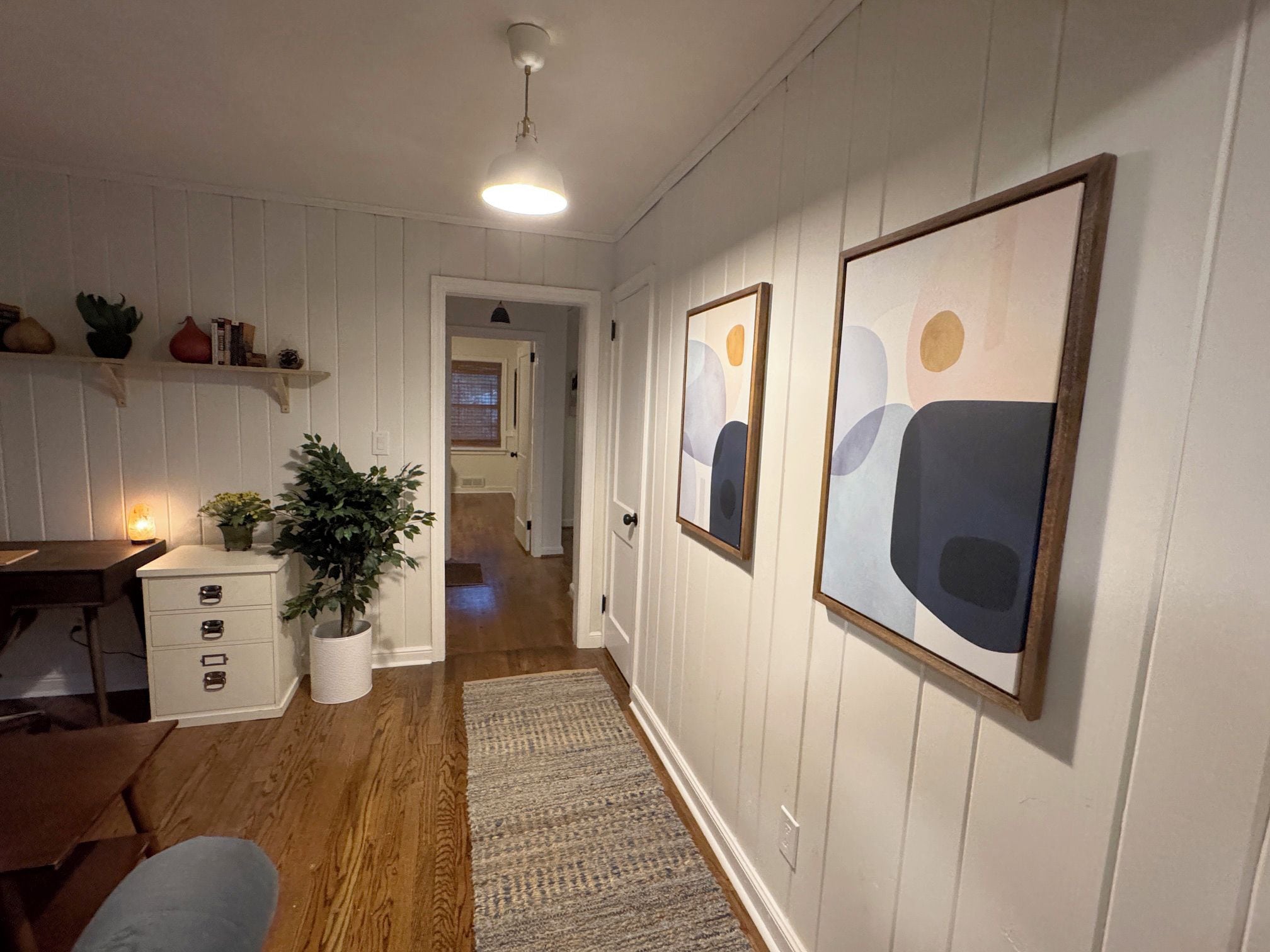Ein Flur mit weißen Wänden, Holzboden, einem Schreibtisch mit Schubladen, einer Zimmerpflanze und abstrakter Kunst an der Wand.