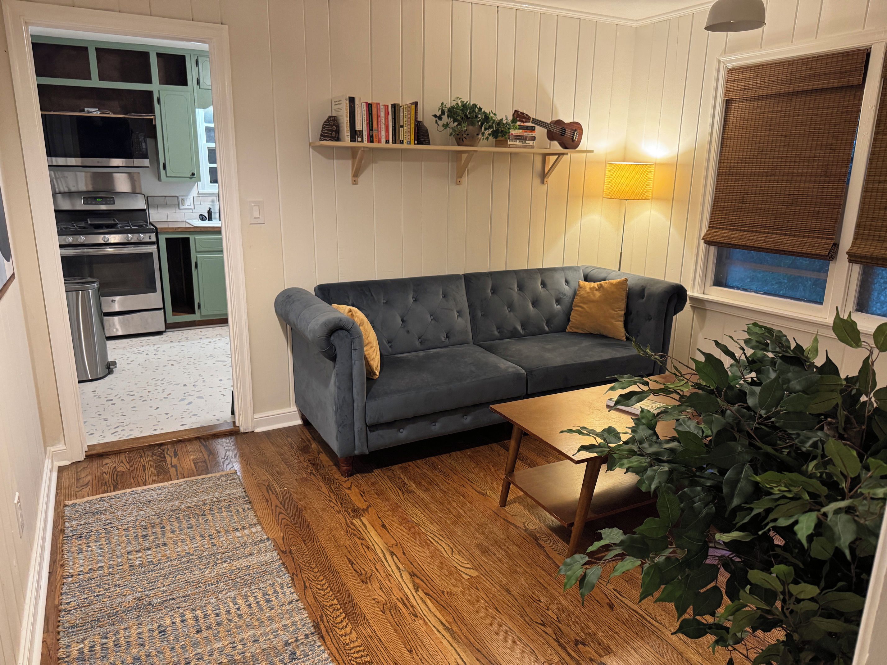 A living room with a dark grey sofa, a wooden coffee table, a patterned rug, a bookshelf with books and a ukulele, and a potted plant.