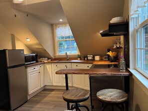 A modern kitchen with white cabinetry, a stainless steel refrigerator, a microwave, and a wooden countertop. There is a window with a view of the outside, and a small table with two stools.