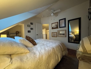 A bedroom with a sloped ceiling, a bed with multiple pillows, a ceiling fan, framed pictures on the wall, and a mirror.