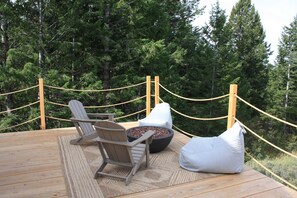 Een houten terras met twee Adirondack-stoelen en een vuurkorf, omringd door een touwbord en dicht bos.