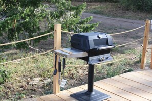 A portable grill on a wooden deck with a rope barrier, surrounded by natural vegetation.