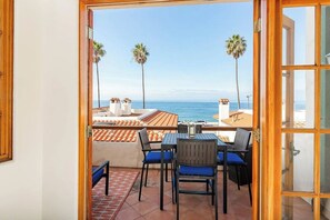 An open door leading to a balcony with a view of the sea, a table with chairs, and palm trees. - Luxury Ocean-View 2-bedroom Condo Across from Beach (Redondo Beach)